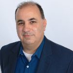 A man with short dark hair wearing a blue shirt and navy blazer poses in front of a plain light background. A man with short dark hair wearing a blue shirt and navy blazer poses in front of a plain light background.