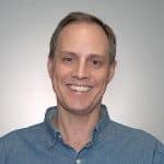 A middle-aged man with short light brown hair, wearing a blue button-up shirt, smiling at the camera against a plain light background.