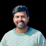 A man with short dark hair and a beard is smiling, wearing a patterned green sweater, against a plain black background. A man with short dark hair and a beard is smiling, wearing a patterned green sweater, against a plain black background.