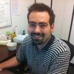 A man with short dark hair and a beard, wearing a black and white striped polo shirt, sits at an office desk and smiles at the camera.