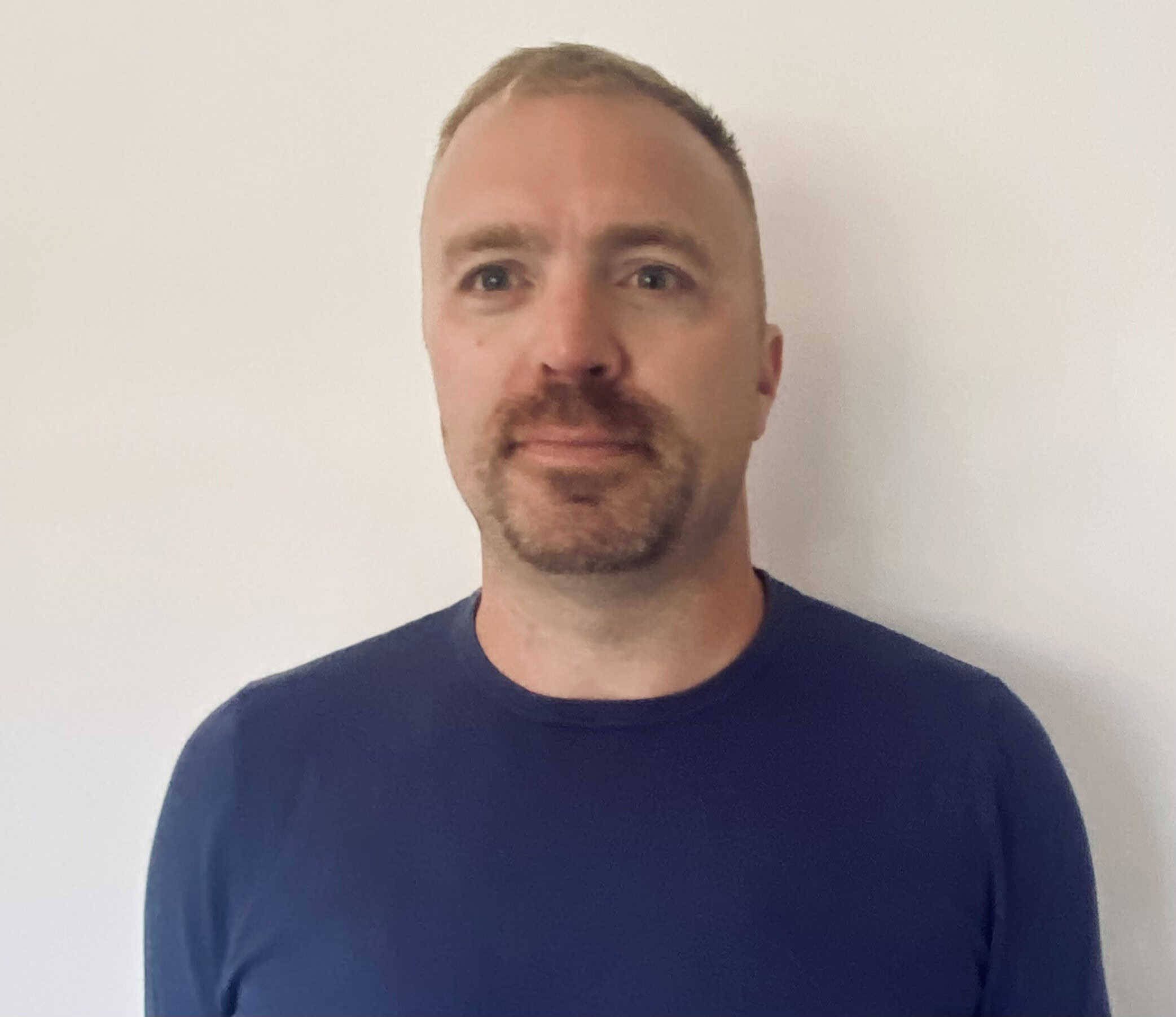 A man with short blond hair, trimmed beard, and mustache, wearing a blue crew-neck shirt, stands in front of a plain white wall, facing the camera.