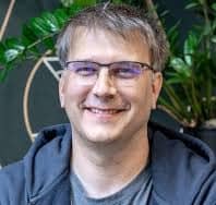 A man with short brown hair and glasses smiles at the camera, wearing a gray shirt and a dark hoodie, with green plants in the background—showcasing Gatling Studio's user-friendly approach to performance testing, where no expertise required.