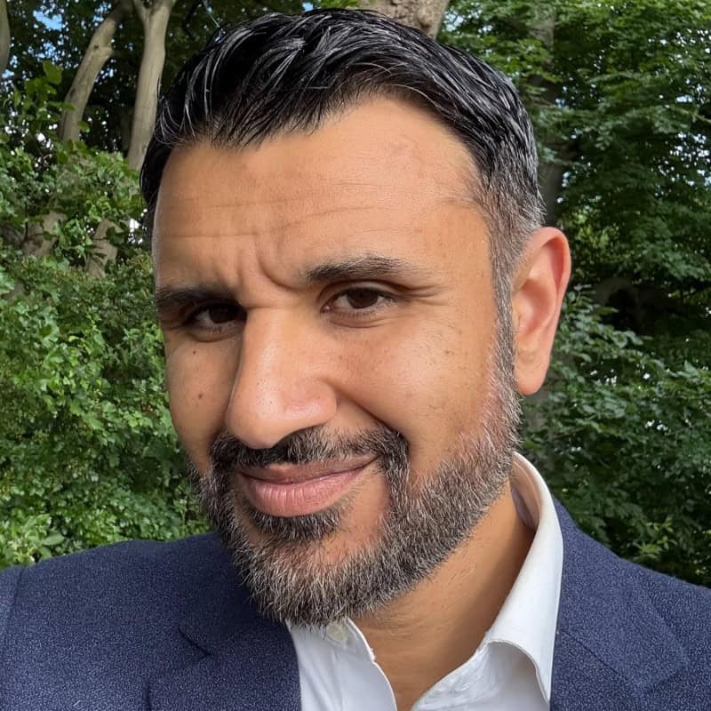 A man with dark hair and a beard, wearing a navy blazer and white shirt, stands outdoors in front of green foliage, thoughtfully discussing LLMs.